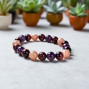 May include: A beaded bracelet featuring a mix of polished gemstones in shades of purple, peach, and burgundy. The bracelet is displayed on a light gray surface, with small potted plants in the background.