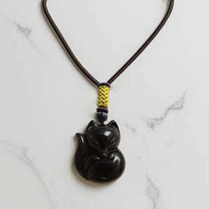 May include: A black fox pendant necklace. The pendant is a stylized fox with a rounded body and a curled tail. The necklace cord is dark brown, with a yellow and dark blue accent near the pendant.