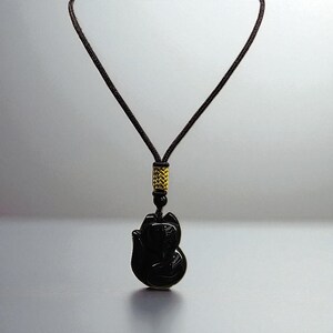 May include: Black fox pendant necklace with a dark brown cord. The pendant is carved in the shape of a fox. The cord has a gold-colored accent near the pendant. The necklace is displayed against a gray background.