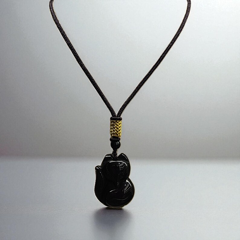 May include: Black fox pendant necklace with a dark brown cord. The pendant is carved in the shape of a fox. The cord has a gold-colored accent near the pendant. The necklace is displayed against a gray background.
