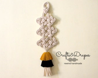 May include: A handmade macrame wall hanging featuring a series of knotted designs in a cream color, topped with a tassel in yellow, black, and cream. The text "Crafts & Drapes" and "rewind handmade" are visible.