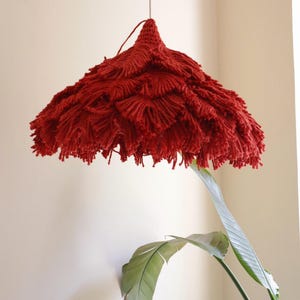 May include: A handmade, red, fringed lampshade hangs from a wire. The lampshade has a conical top and a tiered, textured design. The fringed edges create a soft, layered effect. A green plant with large leaves is in the foreground.
