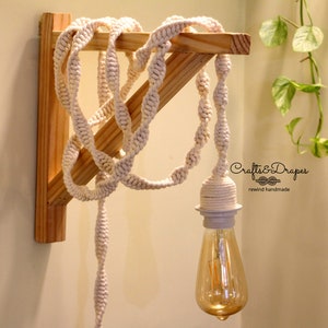 Könnte beinhalten: Eine weiße Baumwollschnur-Leuchte, die an einem Holzregal hängt. Die Schnur ist in einem dekorativen Muster verknotet. Die Glühbirne ist eine klare Edison-Glühbirne aus Glas.