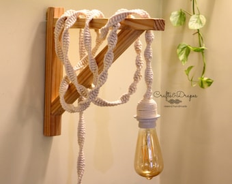 Op de afbeelding: Een witte katoenen snoerlamp die aan een houten plank hangt. Het snoer is in een decoratief patroon geknoopt. De gloeilamp is een heldere glazen Edison-lamp.