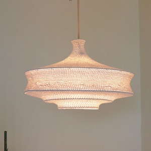 May include: A woven pendant light fixture with a tiered, bell-shaped design. The light is a warm, off-white color and hangs from a thin, brown cord. The fixture is suspended against a neutral-toned wall.
