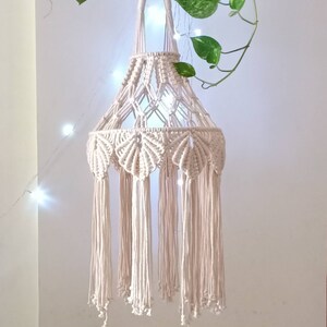 Puede incluir: Una lámpara colgante de macramé blanca con un diseño tejido y flecos. La lámpara tiene un lazo en la parte superior para colgar.