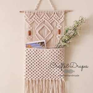 May include: A macrame wall hanging with a pocket for storage. The hanging features a geometric design with wooden beads and a fringe border. The text "Crafts & Drapes rewind handmade" is visible in the bottom right corner.