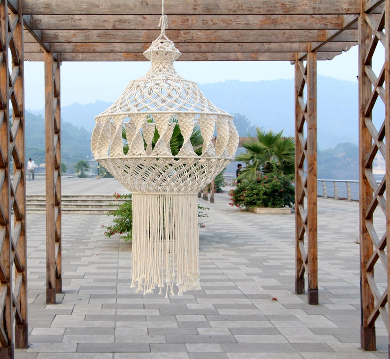 K&ouml;nnte beinhalten: Eine wei&szlig;e Makramee-H&auml;ngelampe mit geometrischem Muster. Die Lampe h&auml;ngt an einer Holzpergola.