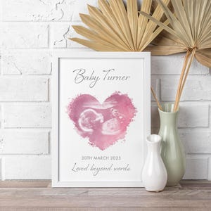 May include: White framed print with the text "Baby Turner" above a pink watercolour heart containing an ultrasound image. The date "20th March 2025" and the phrase "Loved beyond words" are below the heart. Vases in white and green are beside the frame.