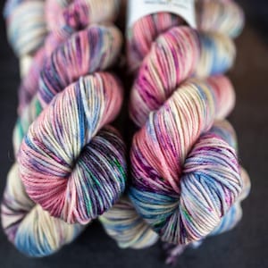 Könnte beinhalten: Nahaufnahme von zwei Strängen bunter Wolle. Die Wolle ist eine Mischung aus Rosa, Lila, Blau und Creme. Die Wolle ist fest gewickelt und wirkt weich. Die Stränge liegen auf einer dunklen Oberfläche. Ideal für Strick- und Häkelprojekte.