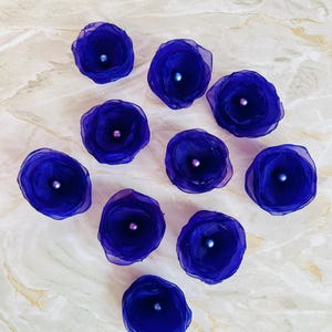 May include: Ten purple fabric flowers with a pearl centre. The flowers are arranged in a semi-circle on a light beige surface.