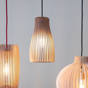 Peut inclure: Trois suspensions en bois de différentes formes, avec une finition naturelle, suspendues sur un fond neutre. Chaque luminaire est composé de lattes verticales en bois créant un effet rayé. Les câbles sont noirs et rouges.