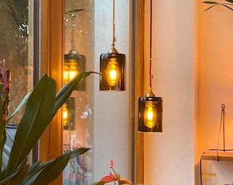 May include: Three glass pendant lights with gold accents hang from the ceiling. The lights are turned on and emit a warm, yellow glow. The lights are hanging in front of a window with a view of a green plant.