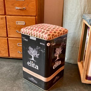 May include: A repurposed stool made from a black olive oil tin. The tin features the brand name "elita" and an olive tree graphic. The cushioned top has a patterned design in shades of pink, red, and brown, with brass-colored tacks.