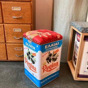 May include: A square olive oil tin repurposed as a stool. The tin is white with blue accents and features the brand name "Regina" and illustrations of olives. The top is upholstered in red fabric with a floral pattern and brass nailhead trim.