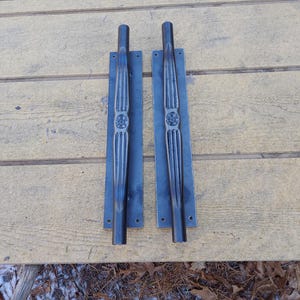 May include: Two dark metal door pulls with a vertical design. Each pull has a rectangular base with mounting holes and a central decorative element. The handles are cylindrical and connected to the base.