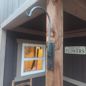 May include: A metal hook attached to a wooden post, designed for hanging items. The hook is curved and mounted on a dark metal plate. A sign that reads "FRESH CUT FLOWERS" is visible.