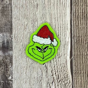 May include: Green felt Grinch character wearing a red and white Santa hat. The Grinch is frowning with his eyes closed.