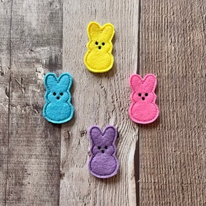May include: Four felt bunny shapes in different colors: yellow, blue, pink, and purple. Each bunny has a simple embroidered face.