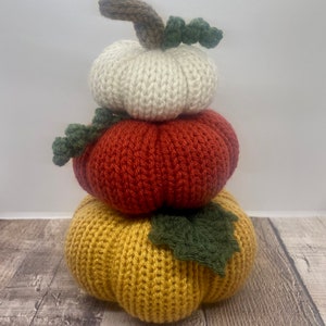 May include: A stack of three knitted pumpkins in fall colors. The top pumpkin is white, the middle pumpkin is orange, and the bottom pumpkin is yellow.