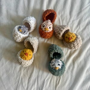 May include: A selection of handmade crochet chicks in eggshell-shaped containers. The chicks are yellow, grey, and brown, with orange beaks. The eggshells are white, beige, and green. The items are arranged on a white surface.