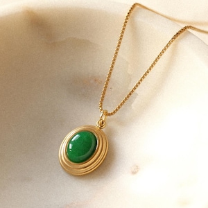 May include: A gold-colored necklace with a green oval-shaped pendant. The pendant is encircled by a series of concentric rings. The necklace is displayed on a white, marble-like surface. The chain is delicate and thin.