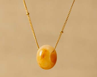 Puede incluir: Un collar de cadena dorada con un único colgante de cuenta ovalada de color ámbar. La cadena presenta pequeñas cuentas doradas espaciadas uniformemente a lo largo de su longitud. El texto AMIOA JEWELS aparece en la esquina inferior derecha.