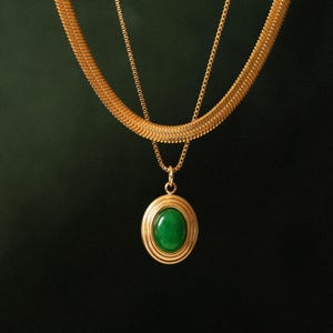 May include: A gold-toned necklace with a green oval pendant. The pendant is set in a gold frame with concentric circles. The necklace has a thick, textured chain and a delicate chain. The background is a dark green.