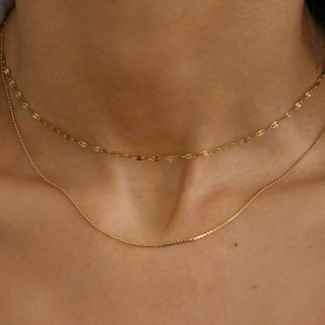 May include: A gold jewellery set comprising thick hoop earrings and two delicate necklaces. The necklaces include a chain with small, flat links and a simple, thin chain. The jewellery is set against a neutral background.