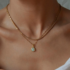 May include: A layered gold-tone necklace set featuring a shorter rope chain and a longer delicate chain with a single teardrop-shaped pendant. The pendant has a smooth, pale iridescent stone set in a gold-coloured metal frame, resting against the collarbone.