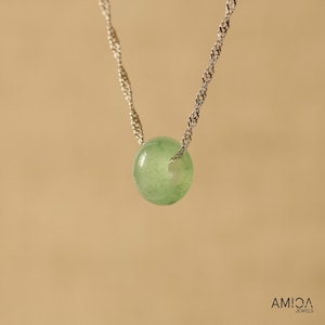May include: A delicate necklace featuring a silver chain suspending a round, pale green gemstone pendant. The pendant is a smooth, polished sphere. The background is a solid, warm beige.