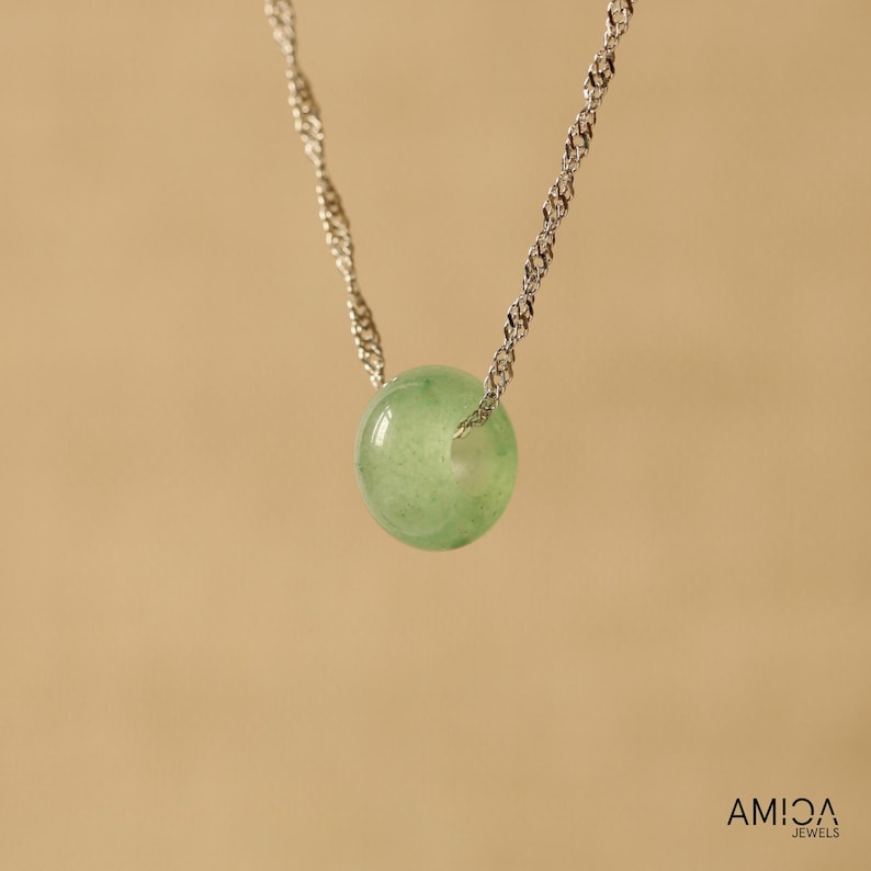 May include: A delicate necklace featuring a silver chain suspending a round, pale green gemstone pendant. The pendant is a smooth, polished sphere. The background is a solid, warm beige.