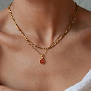 May include: A layered gold necklace with a teardrop pendant. The necklace has two chains: a rope chain and a delicate chain. The pendant is a vibrant red-orange colour. The jewellery is worn around the neck.
