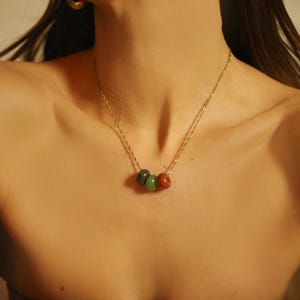 May include: A gold-toned necklace featuring three gemstone beads in shades of green and red. The necklace is worn against a brown strapless top. Gold hoop earrings are also visible.