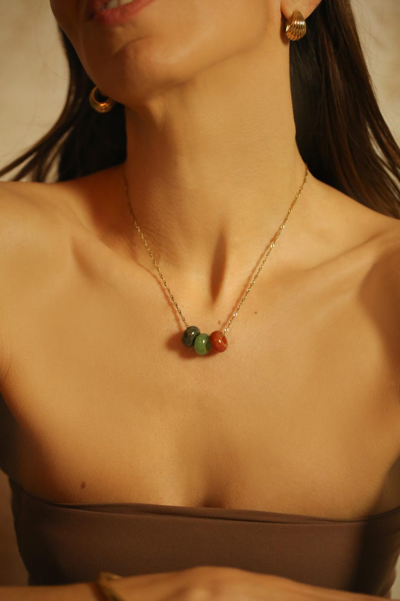 May include: A gold-toned necklace featuring three gemstone beads in shades of green and red. The necklace is worn against a brown strapless top. Gold hoop earrings are also visible.