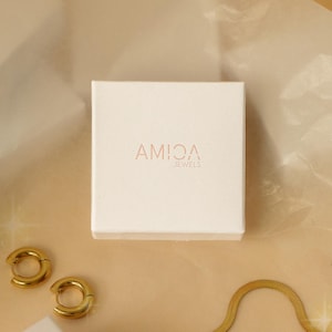 May include: A square, off-white jewelry box with the brand name "AMICA JEWELS" in a light brown font. Two gold hoop earrings and a gold chain are visible. The background is a light brown, textured surface.