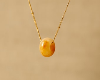 Puede incluir: Un collar de cadena dorada con un único colgante de cuenta ovalada de color ámbar. La cadena presenta pequeñas cuentas doradas espaciadas uniformemente a lo largo de su longitud. El texto AMIOA JEWELS aparece en la esquina inferior derecha.
