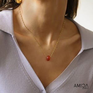 May include: A delicate gold necklace features a single, small, reddish-orange gemstone pendant. The chain is thin and delicate, and the gemstone appears smooth and subtly textured.  The necklace is displayed against a light purple background. The brand name AMICA JEWELS is visible in the bottom right corner.