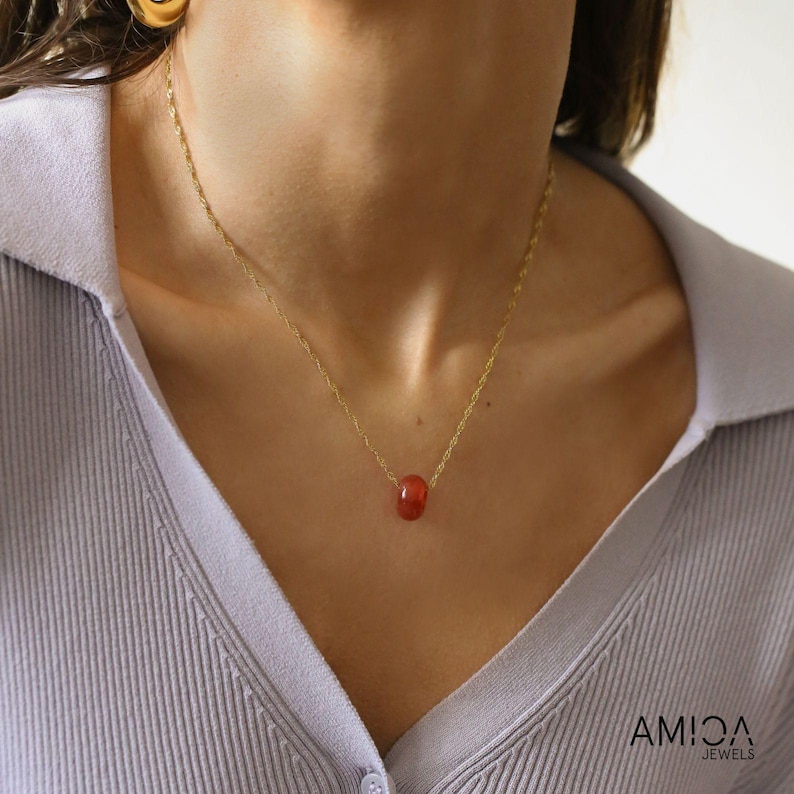 May include: A delicate gold necklace features a single, small, reddish-orange gemstone pendant. The chain is thin and delicate, and the gemstone appears smooth and subtly textured.  The necklace is displayed against a light purple background. The brand name AMICA JEWELS is visible in the bottom right corner.