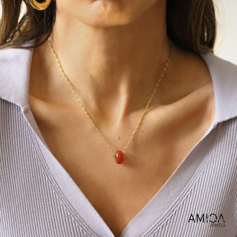 May include: A gold chain necklace with a small, cylindrical, reddish-orange gemstone pendant. The necklace is worn against a light blue collared shirt. The brand name "AMIDA JEWELS" is visible in the bottom right corner.