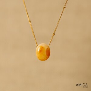 May include: A gold-colored necklace featuring a large, oval-shaped, amber-colored stone pendant. The pendant is suspended from a delicate gold chain. The necklace is displayed against a neutral background. The brand name "AMICA JEWELS" is visible in the bottom right corner.