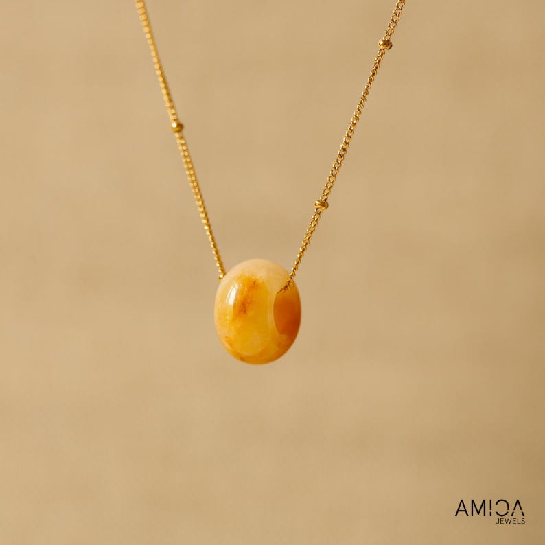 May include: A gold-colored necklace featuring a large, oval-shaped, amber-colored stone pendant. The pendant is suspended from a delicate gold chain. The necklace is displayed against a neutral background. The brand name "AMICA JEWELS" is visible in the bottom right corner.