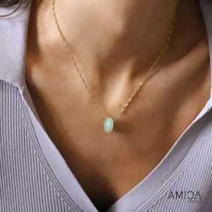 May include: A delicate gold necklace featuring a light green, oval-shaped gemstone pendant. The necklace is worn against a light purple shirt. The jewelry is from Amica Jewels.
