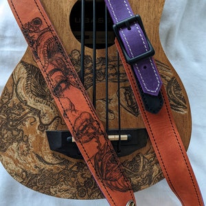 Może przedstawiać: Dwa sk&oacute;rzane paski do gitary, jeden pomarańczowy z czarno-białym wzorem smoka i jeden fioletowy z białymi szwami. Paski leżą na ukulele.