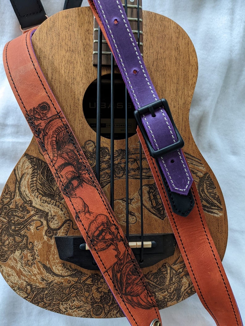 Może przedstawiać: Dwa sk&oacute;rzane paski do gitary, jeden pomarańczowy z czarno-białym wzorem smoka i jeden fioletowy z białymi szwami. Paski leżą na ukulele.