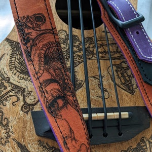 Może przedstawiać: Ukulele z brązowym drewnianym korpusem i czarno-białym wzorem. Ukulele ma czarny mostek i zestaw strun. Do ukulele przymocowane są dwa sk&oacute;rzane paski do gitary. Jeden pasek jest brązowy z fioletowym brzegiem i czarno-białym wzorem. Drugi pasek jest fioletowy z białymi szwami i czarną klamrą.