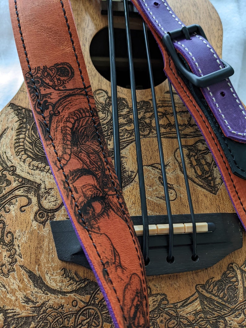 Może przedstawiać: Ukulele z brązowym drewnianym korpusem i czarno-białym wzorem. Ukulele ma czarny mostek i zestaw strun. Do ukulele przymocowane są dwa sk&oacute;rzane paski do gitary. Jeden pasek jest brązowy z fioletowym brzegiem i czarno-białym wzorem. Drugi pasek jest fioletowy z białymi szwami i czarną klamrą.