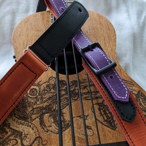 Może przedstawiać: Ukulele z brązowym drewnianym korpusem i czarno-białym wzorem. Dwa paski do gitary są owinięte wok&oacute;ł ukulele. Jeden pasek jest fioletowy z białymi szwami, a drugi jest pomarańczowy z brązowymi szwami.