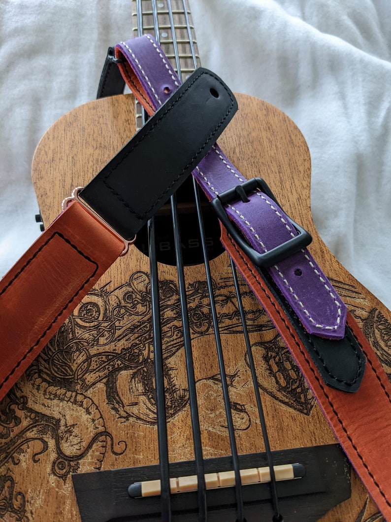 Może przedstawiać: Ukulele z brązowym drewnianym korpusem i czarno-białym wzorem. Dwa paski do gitary są owinięte wok&oacute;ł ukulele. Jeden pasek jest fioletowy z białymi szwami, a drugi jest pomarańczowy z brązowymi szwami.