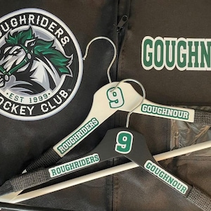 May include: Two wooden clothes hangers, one white and one black, with the number 9 and the name "GOUGHNOUR" printed on them. The hangers also have the text "ROUGHRIDERS" printed on them. A circular logo for the "ROUGHRIDERS HOCKEY CLUB" is also visible.